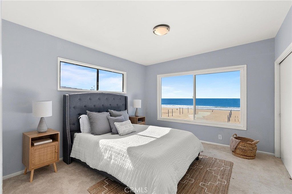 This bright bedroom features a dark upholstered headboard and a neatly made bed with white bedding, flanked by two light wood nightstands with matching lamps. A large window provides a stunning, unobstructed view of the beach and ocean, creating a serene and airy atmosphere. The room is finished with light blue walls and a textured area rug over neutral carpeting, offering a clean and coastal aesthetic.