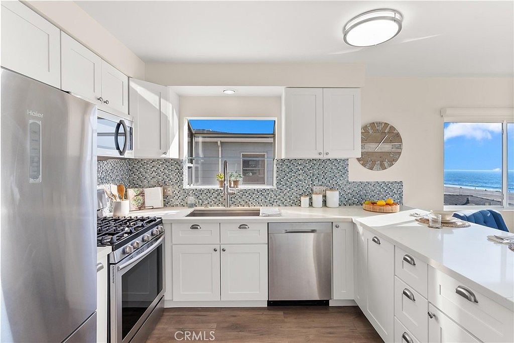 This bright, modern kitchen features crisp white cabinetry, stainless steel appliances, and a striking blue and white mosaic tile backsplash. A peninsula with a white countertop extends into the space, providing additional workspace and a view of the ocean through a large window. The room is illuminated by recessed lighting and offers a clean, coastal-inspired aesthetic.