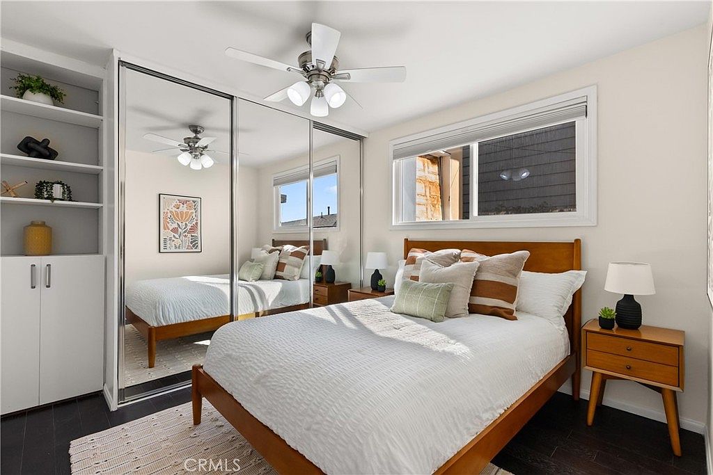 This cozy bedroom features a mid-century modern aesthetic with a warm wood-framed bed and matching nightstand. A large mirrored closet door reflects the room, creating a sense of depth, while a ceiling fan with integrated lighting provides overhead illumination. The space is accented by a textured rug, neutral bedding, and a framed piece of wall art, offering a clean and inviting atmosphere.