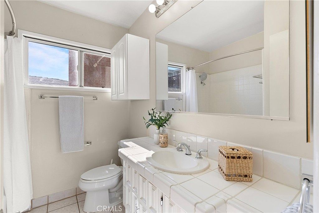 This bright bathroom features a white tiled vanity countertop with an integrated sink and a large, wall-to-wall mirror that enhances the sense of space. A white toilet is positioned next to the vanity, and a shower-tub combination with a white curtain is visible in the background. The room is illuminated by a multi-bulb vanity light fixture above the mirror and natural light streaming through a window above the toilet.
