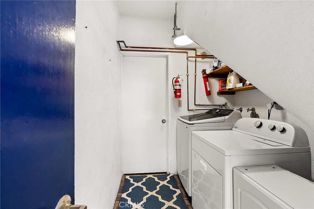 This functional laundry room features a side-by-side washer and dryer set positioned under a sloped ceiling. The space includes a white door, exposed copper plumbing pipes along the wall, and a small wooden shelf holding cleaning supplies and a fire extinguisher. A blue and white patterned rug adds a touch of color to the floor, creating a compact and utilitarian laundry area.