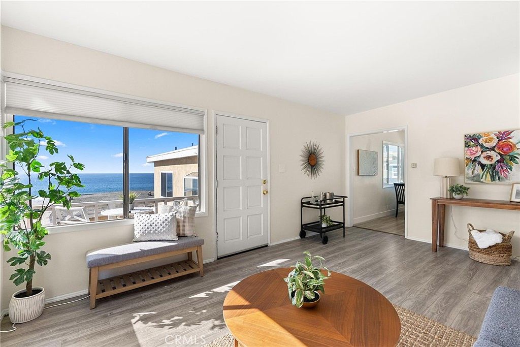 This bright living room features light-toned walls and wood-look flooring, creating an airy and inviting atmosphere. A large window provides a stunning, direct view of the ocean and beach, while a wooden bench with decorative pillows sits beneath it. The space is furnished with a round wooden coffee table, a bar cart, and a console table, all arranged to highlight the open layout and coastal aesthetic.