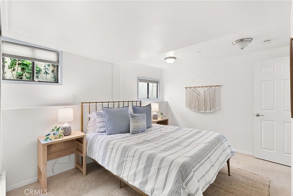This bright and airy bedroom features a neutral color palette with a queen-sized bed dressed in blue and white striped bedding. The room is accented by a decorative macramé wall hanging, two light wood nightstands with matching table lamps, and two windows that allow for natural light. The space has a clean, minimalist aesthetic with light-colored carpeting and white walls, creating a calm and inviting atmosphere.