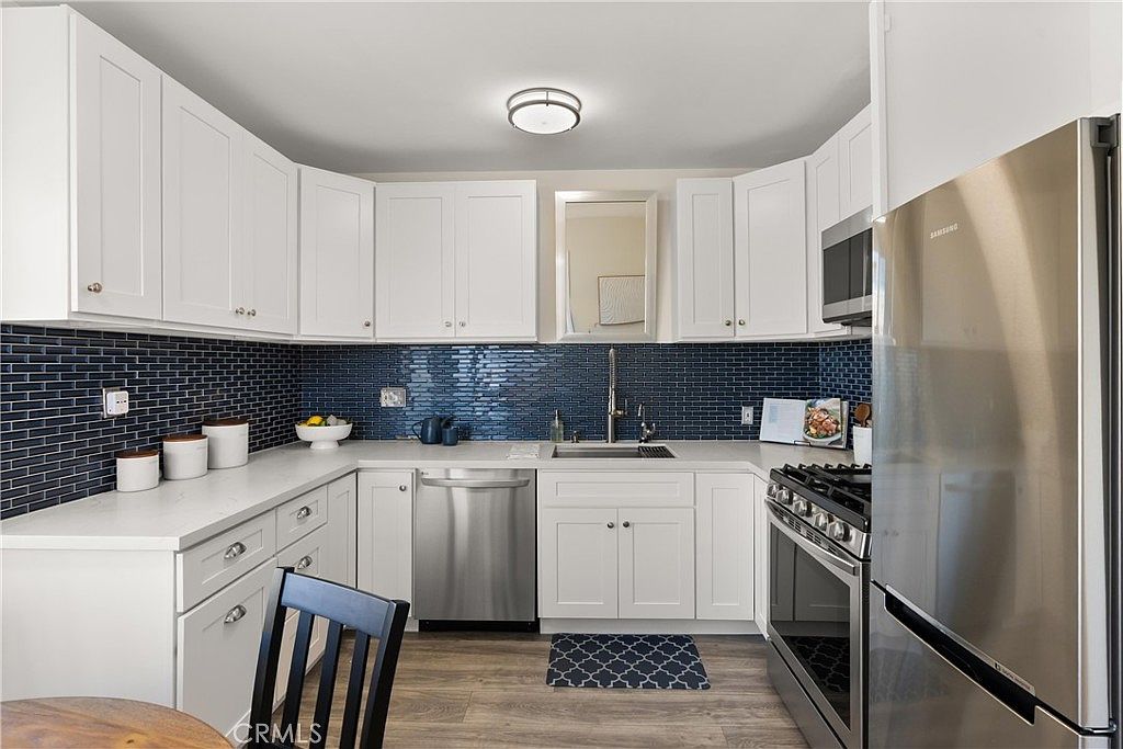 This bright, U-shaped kitchen features crisp white cabinetry paired with a striking dark blue subway tile backsplash. The space is equipped with stainless steel appliances, including a gas range and a dishwasher, set against light-toned countertops. A wood-look floor runs throughout the area, creating a clean and modern aesthetic that feels both functional and inviting.
