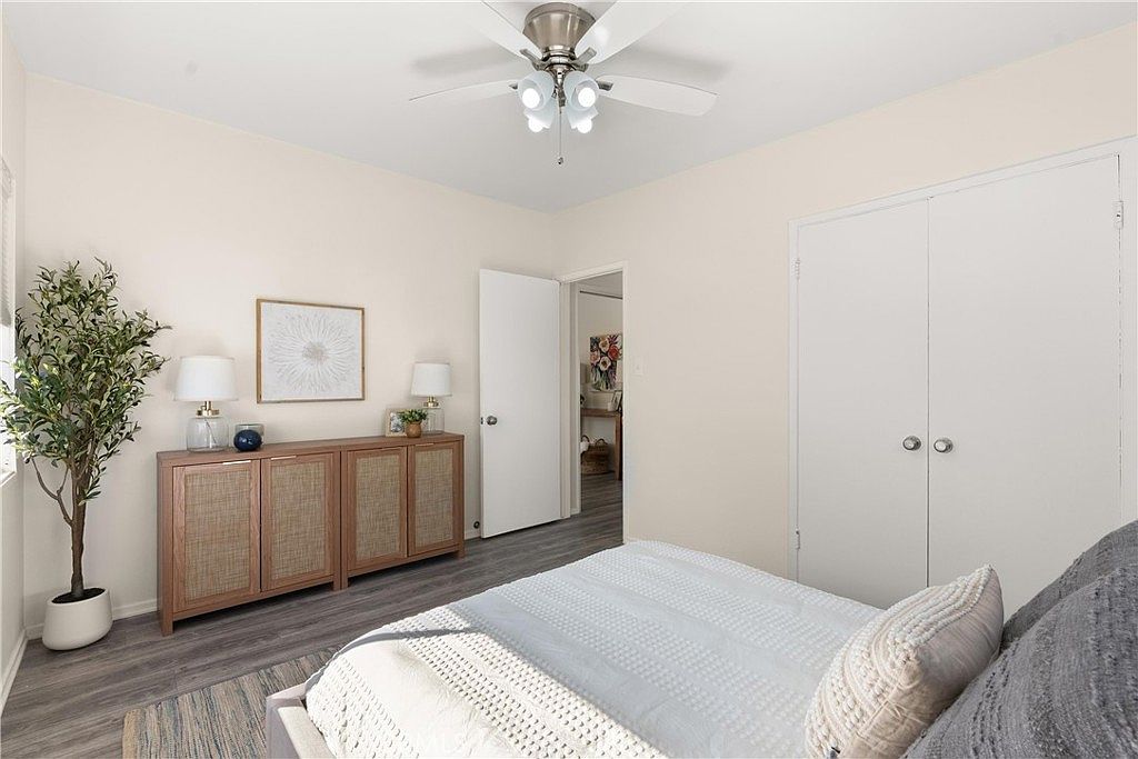 This bright and neutral bedroom features light-toned walls, wood-look flooring, and a ceiling fan with integrated lighting. A wooden console with woven cabinet doors sits against the wall, topped with two lamps and a framed piece of art, while a bed with a textured gray comforter occupies the foreground. The room includes a white closet with double doors and an open doorway leading to an adjacent space, creating an airy and inviting atmosphere.