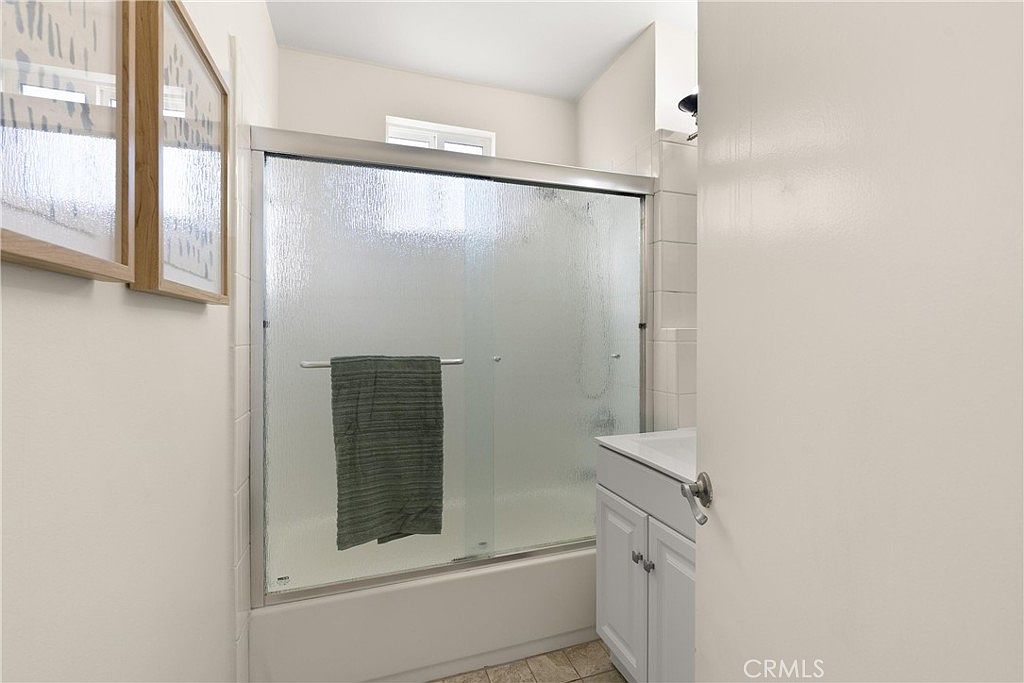 This bathroom features a white vanity with a sink and a shower-tub combination enclosed by a sliding glass door. The walls are finished with white tiles, and a dark towel hangs over the shower door. The perspective is from the doorway, looking into the compact, functional space.