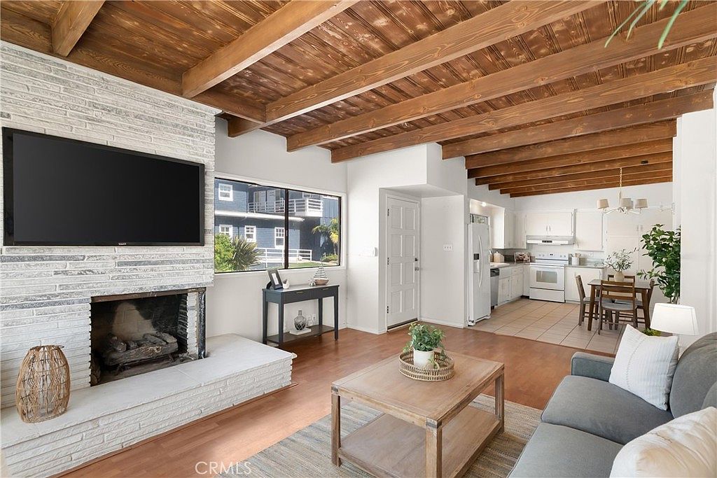 This open-concept living area features a prominent white-painted brick chimney breast with a mounted television above the fireplace. The space is defined by exposed wood ceiling beams and warm-toned wood flooring, creating a cozy, rustic atmosphere. A neutral-toned sofa and a wooden coffee table anchor the seating area, which flows seamlessly into a bright kitchen and dining space in the background.