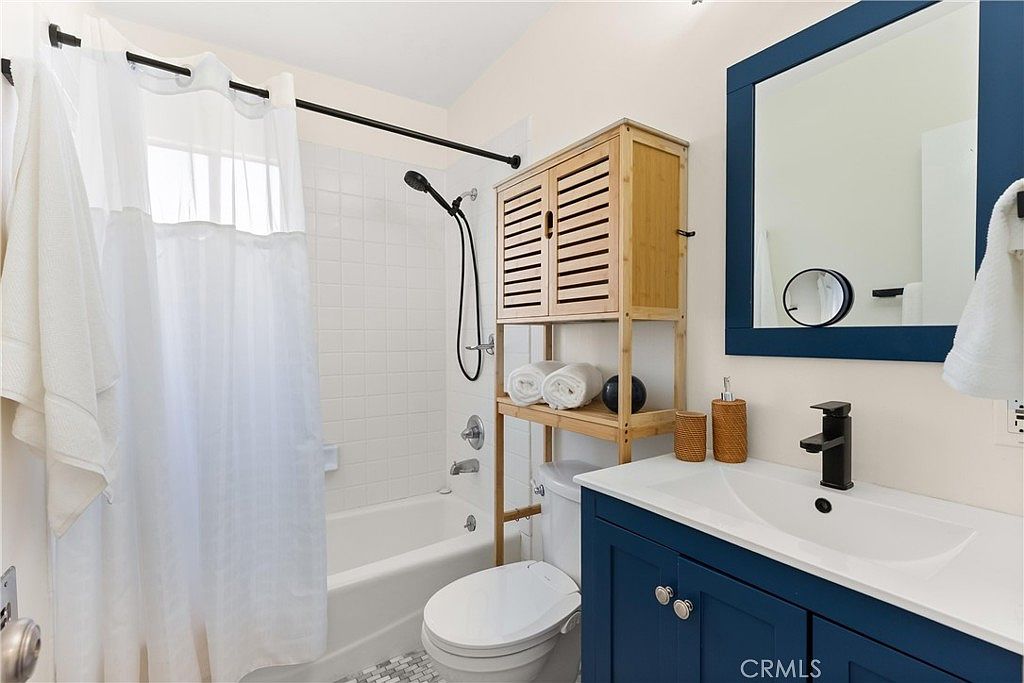 This bright and compact bathroom features a navy blue vanity with a modern black faucet and a rectangular sink. A white shower-tub combination with a white curtain is positioned next to a toilet, which is accented by a light wood shelving unit holding rolled towels. The space is finished with a framed mirror and a patterned tile floor, creating a clean and functional aesthetic.