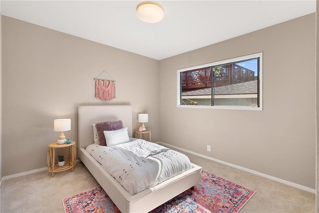 This bright and neutral guest bedroom features a light-colored upholstered bed frame centered against a soft beige wall. A patterned, colorful area rug adds a vibrant touch to the carpeted floor, while a window provides natural light and a view of an outdoor deck. The room is accented with matching bedside tables, lamps, and a decorative wall hanging, creating a cozy and inviting atmosphere.