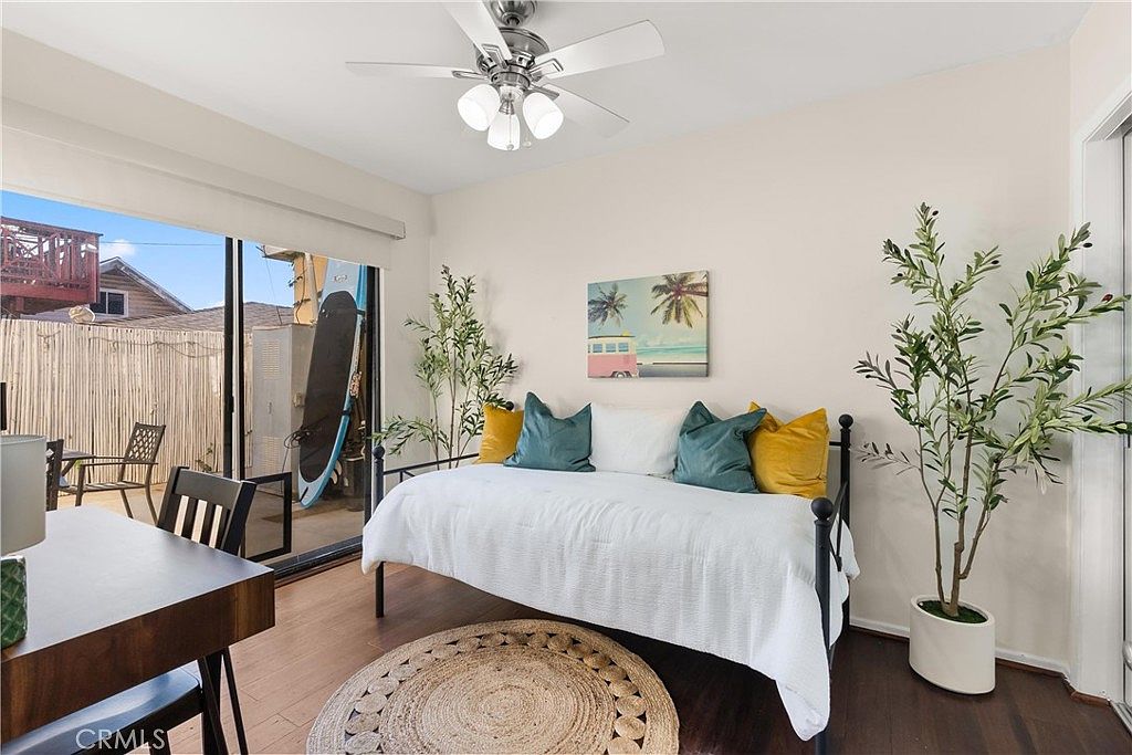 This bright guest bedroom features a daybed styled with colorful throw pillows and a neutral white quilt, centered against a light-toned wall. A round jute rug sits on the dark wood flooring, while a potted plant adds a touch of greenery to the corner. Large sliding glass doors provide access to a private patio area, allowing for ample natural light to fill the space.