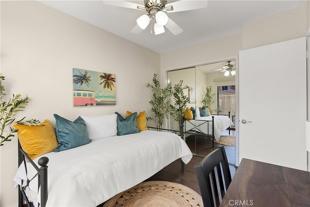 This guest bedroom features a daybed styled with teal and mustard yellow pillows, positioned against a light-toned wall adorned with a beach-themed canvas print. A large mirrored closet door reflects the room, creating a sense of depth and showing a glimpse of a bed in the reflection. The space is accented by potted greenery and a round woven rug, offering a bright and inviting atmosphere.