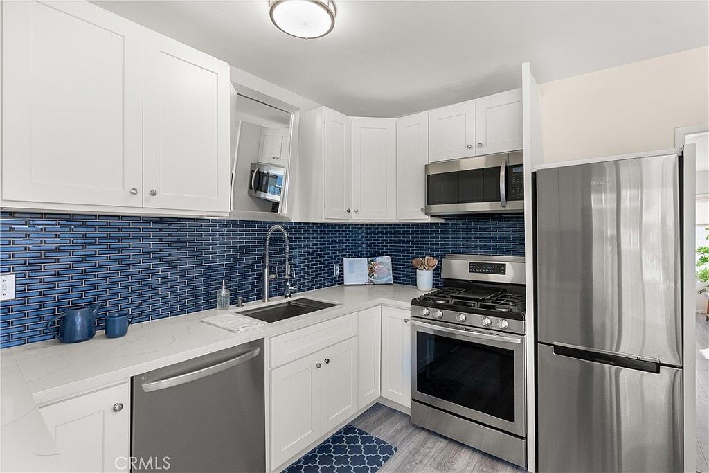 This bright, modern kitchen features crisp white cabinetry paired with a vibrant blue subway tile backsplash. The space is equipped with stainless steel appliances, including a gas range and a refrigerator, set against light-colored countertops. The perspective is a straight-on eye-level shot that highlights the efficient U-shaped layout and clean, contemporary aesthetic.