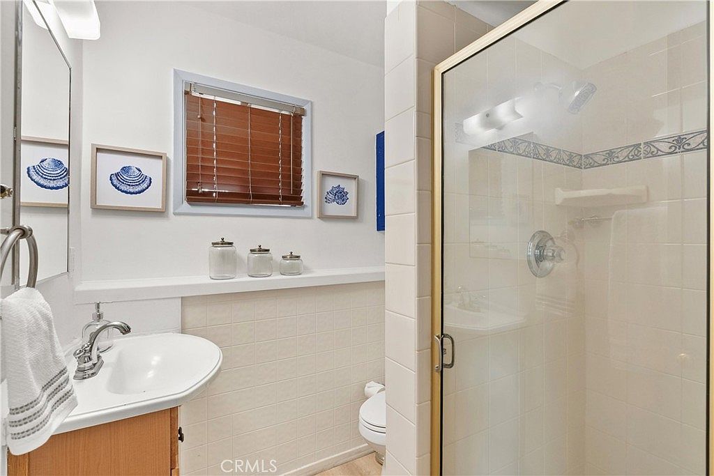 This compact bathroom features a pedestal sink, a toilet, and a glass-enclosed shower stall with tiled walls. The space is decorated with nautical-themed artwork and a wooden window blind, creating a clean and functional aesthetic. The perspective captures the entire room from the doorway, highlighting the efficient layout and neutral color palette.