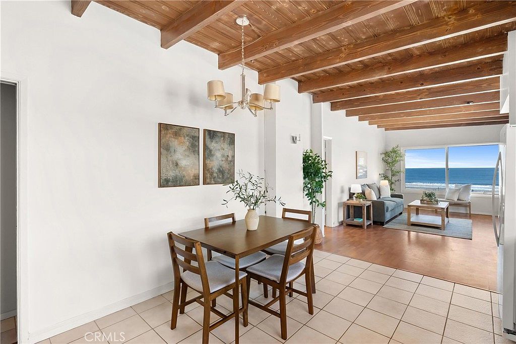 This open-concept dining area features a wooden table with four chairs, positioned beneath a classic chandelier with exposed wooden beams on the ceiling. The space flows seamlessly into a living area that showcases a stunning, unobstructed view of the ocean through large windows. The interior design combines a rustic, coastal aesthetic with neutral walls and light-colored tile flooring, creating a bright and airy atmosphere.