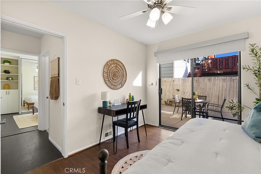 This bedroom features a neutral color palette with dark wood-style flooring and a sliding glass door that opens to a private patio. A small desk area is positioned against the wall, accented by a decorative woven wall hanging, while a ceiling fan provides overhead lighting. The room offers a view into an adjacent hallway with built-in shelving, creating a functional and well-lit living space.