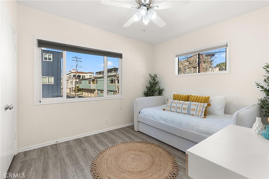 This bright and airy bedroom features light-toned walls, wood-look flooring, and a round woven area rug that adds texture to the space. A daybed with decorative pillows is positioned against the wall, complemented by a white desk and a ceiling fan with integrated lighting. Large windows provide ample natural light and a view of the neighboring buildings, creating a clean and inviting atmosphere.
