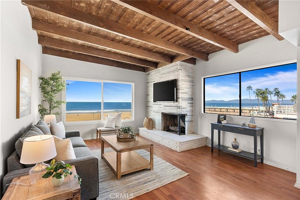 This bright living room features a vaulted ceiling with exposed wooden beams and warm-toned wood flooring. A prominent stone-clad chimney breast serves as a focal point, complemented by large windows that offer expansive views of the beach and ocean. The space is furnished with a comfortable gray sofa, a light wood coffee table, and a console table, creating an inviting and airy coastal atmosphere.