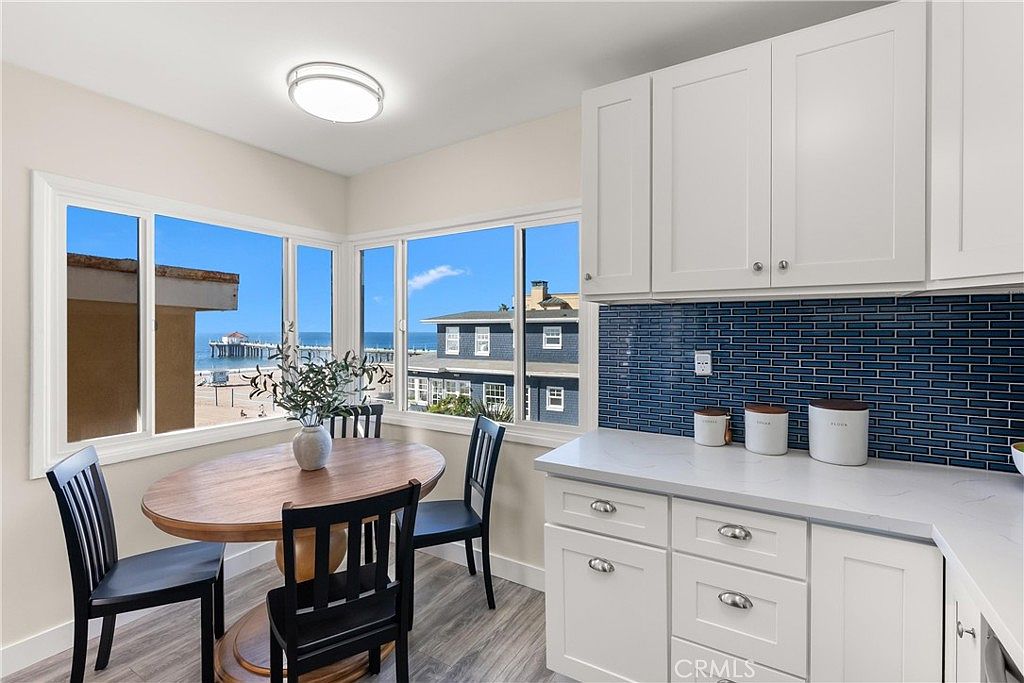 This bright kitchen features white cabinetry paired with a striking blue tiled backsplash and light-toned countertops. A cozy dining area is positioned by large windows that offer a scenic view of the ocean and a nearby pier. The space combines functional cooking areas with a casual breakfast nook, creating an inviting and airy atmosphere.