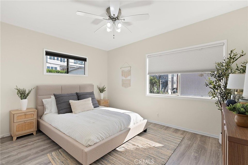 This bright and airy bedroom features light-toned walls and wood-look flooring, creating a clean and modern aesthetic. The room is centered around a bed with a neutral upholstered headboard, flanked by two matching light wood nightstands topped with small potted plants. Large windows with blinds allow natural light to fill the space, while a ceiling fan provides overhead lighting and ventilation.