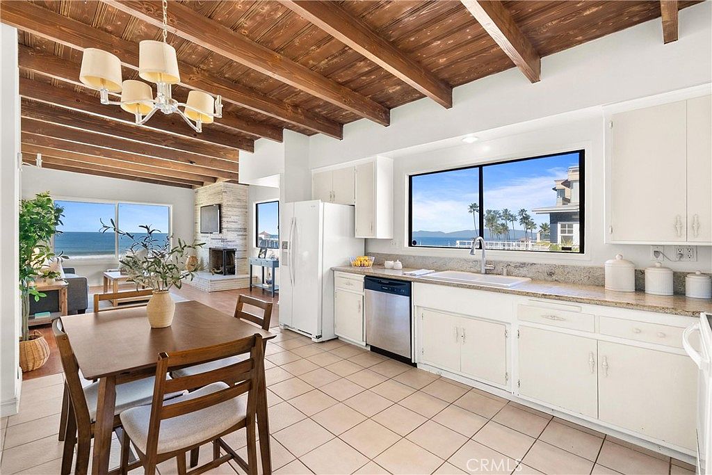 This open-concept kitchen features white cabinetry, light-toned stone countertops, and a dining area with a wooden table. The space is defined by a rustic wood-beamed ceiling and large windows that provide a clear view of the ocean and coastline. The perspective captures the flow from the kitchen into the adjacent living area, which features a stone-clad chimney breast, creating a bright and airy coastal atmosphere.
