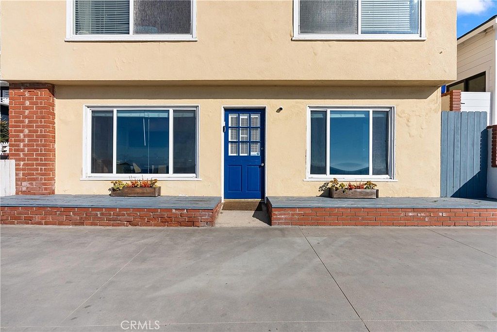 This front-facing view of a two-story residence features a light-colored stucco facade with a prominent blue front door centered between two large windows. The ground level includes brick-accented planters and a concrete driveway, while the upper level showcases two additional windows. The architectural style is simple and functional, with a clean, symmetrical layout that provides a clear view of the home's entrance.