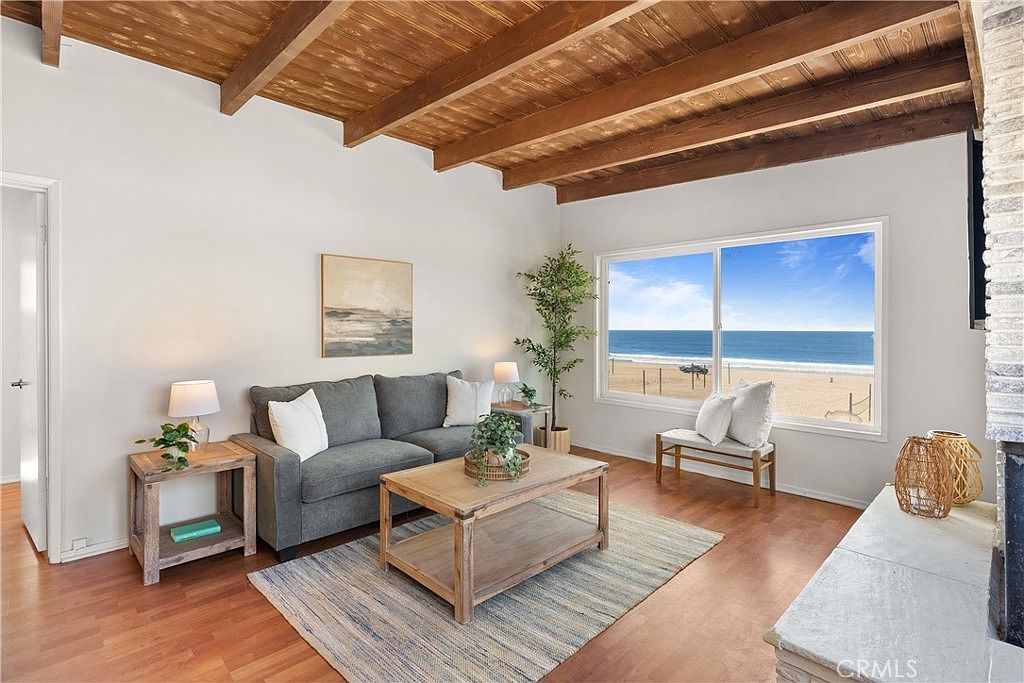 This bright living room features a vaulted ceiling with exposed wood beams and warm wood-toned flooring. A comfortable gray sofa is positioned in front of a large window that provides a direct, unobstructed view of the beach and ocean. The space is decorated with a light wood coffee table, a textured area rug, and a tall indoor plant, creating a relaxed, coastal-inspired atmosphere.