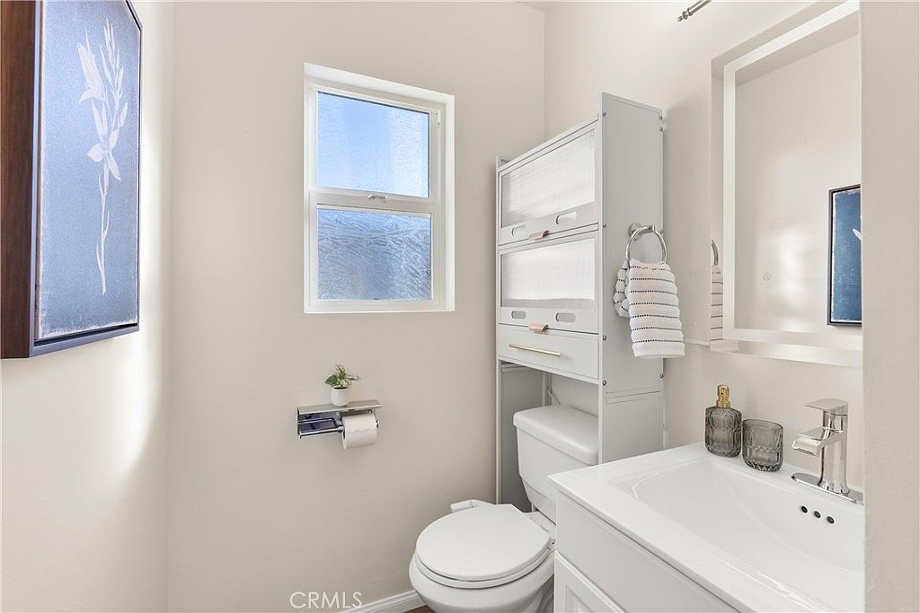 This compact bathroom features a white vanity with a rectangular sink, a toilet, and a tall, multi-tiered storage unit positioned behind the toilet. The walls are painted a neutral tone, complemented by a window that provides natural light and framed artwork on the wall. The space is organized and functional, presenting a clean and tidy aesthetic suitable for a guest powder room.