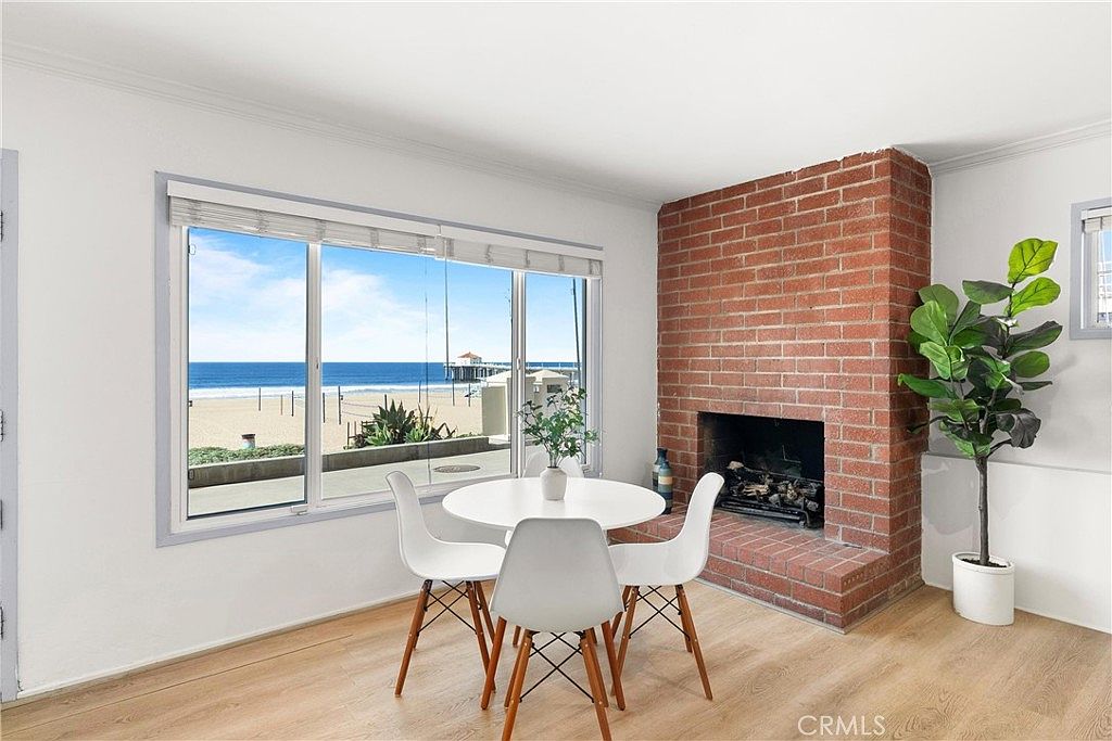 This bright dining area features a round white table with three modern chairs, positioned perfectly to take advantage of the expansive ocean and pier views through large windows. To the right, a prominent red brick chimney breast adds a rustic, textured element to the otherwise minimalist white-walled space. A potted plant stands beside the brick feature, and the room is finished with light wood-toned flooring, creating a serene and airy coastal atmosphere.