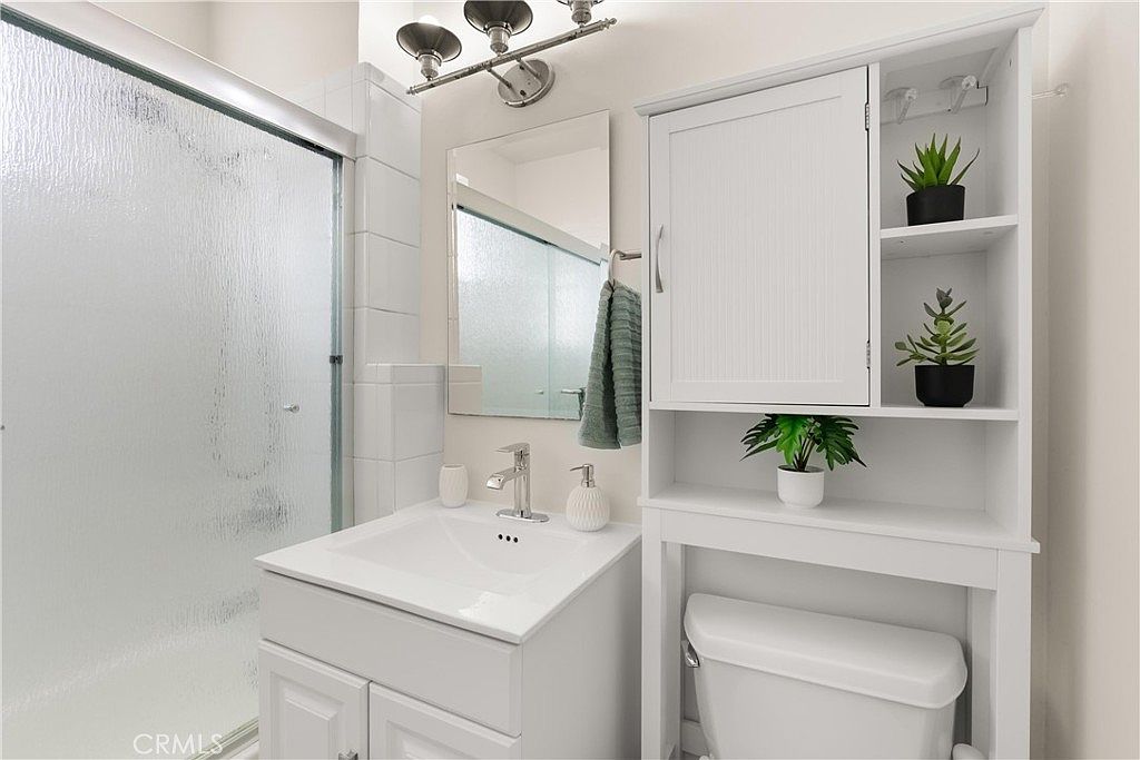 This compact, modern bathroom features a white vanity with a rectangular sink and a matching over-the-toilet storage cabinet. A glass-enclosed shower stall sits to the left, while a wall-mounted mirror and a three-light vanity fixture provide illumination. The space is bright and functional, utilizing vertical storage to maximize the footprint of the room.