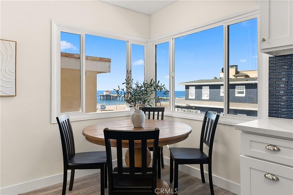 This bright dining area features a round wooden table surrounded by three black chairs, positioned perfectly to take advantage of the expansive corner windows. The windows offer a stunning, unobstructed view of the ocean, beach, and a nearby pier, creating a serene and airy atmosphere. A simple vase with greenery sits as a centerpiece on the table, complementing the clean, minimalist aesthetic of the space.