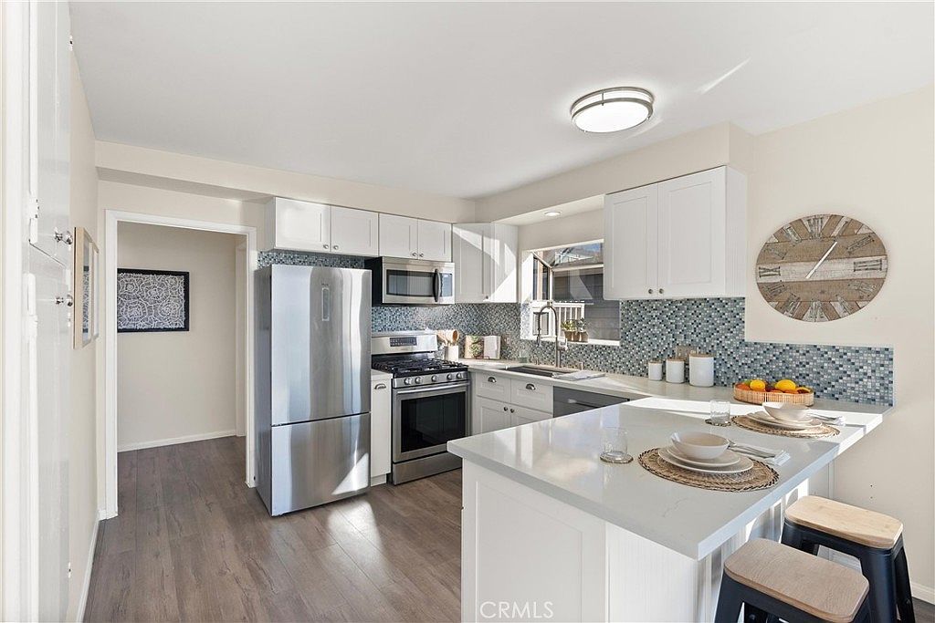 This bright, modern kitchen features white cabinetry, stainless steel appliances, and a peninsula with a light-toned countertop that provides additional seating. A distinctive blue and white mosaic tile backsplash adds a pop of color, while a large wall clock serves as a focal point on the side wall. The space is illuminated by a flush-mount ceiling light and natural light from the window above the sink, creating an open and inviting atmosphere.