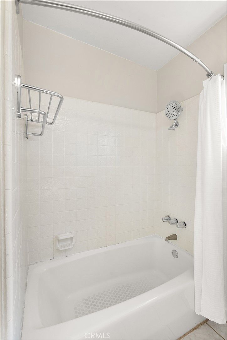 This bathroom features a standard white bathtub with a tiled surround and a shower head. A chrome towel rack is mounted on the left wall, and a white shower curtain hangs from a curved rod. The space is finished in neutral tones, providing a clean and functional aesthetic.