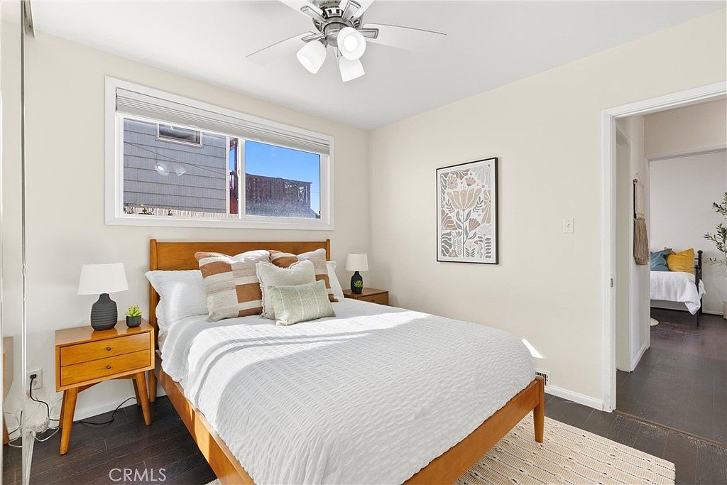 This bright and inviting bedroom features a wooden bed frame centered against a neutral-toned wall, accented by a framed floral art piece. A large window allows natural light to fill the room, highlighting the dark wood-look flooring and the cozy bedding. The space is well-appointed with a matching wooden nightstand and a ceiling fan, creating a comfortable and clean atmosphere.