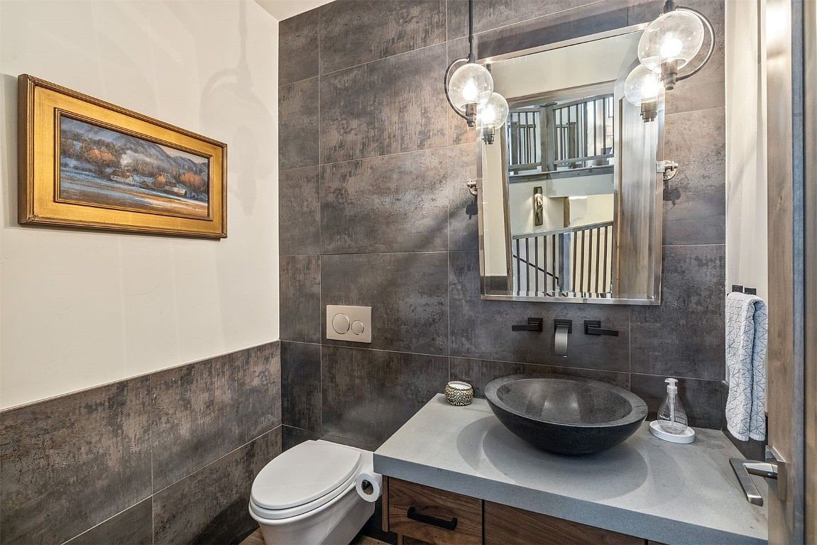 This is a stylish guest bathroom featuring a modern design with gray tiled walls and a half wall of white paint. A vessel sink sits atop a wooden vanity with a light gray countertop, complemented by a sleek, wall-mounted faucet. The room is illuminated by a unique pendant light fixture, and a framed painting adds a touch of color to the space.