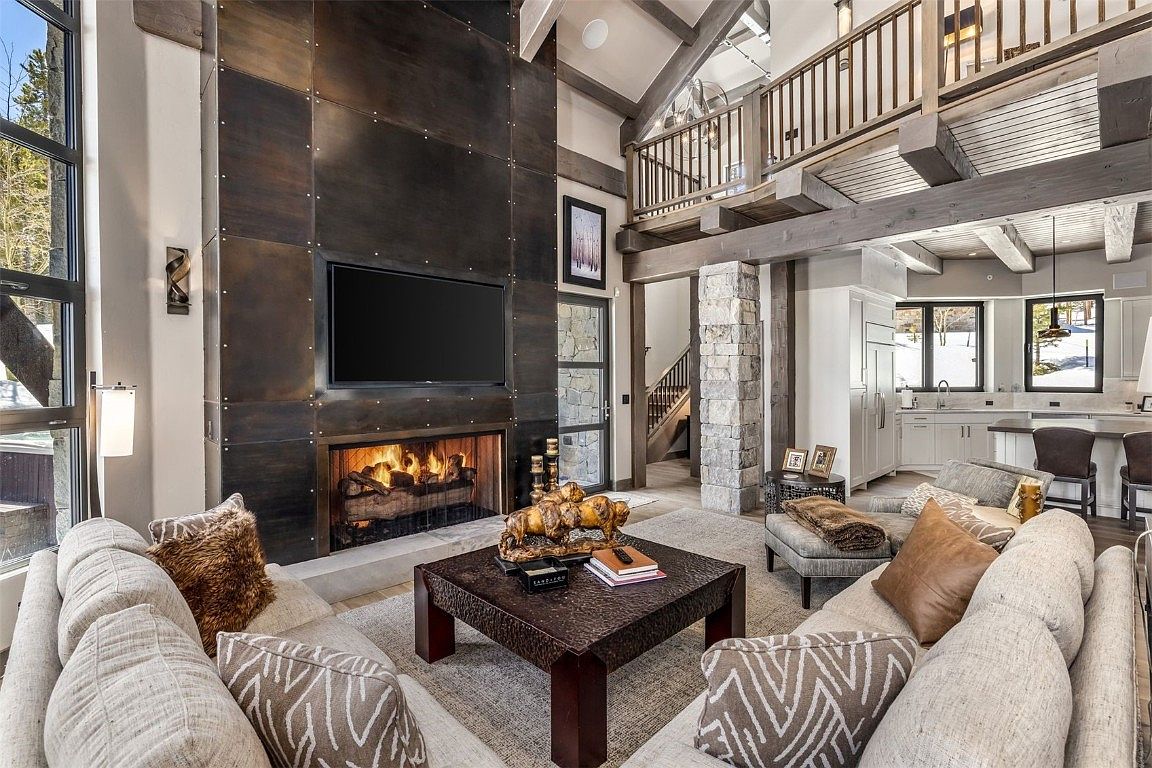 This is a spacious living room featuring a floor-to-ceiling metal-clad fireplace with a mounted television above. The room is furnished with a large sectional sofa, a square coffee table, and an area rug. Stone pillars and exposed beams add to the rustic-modern aesthetic, while large windows provide natural light.