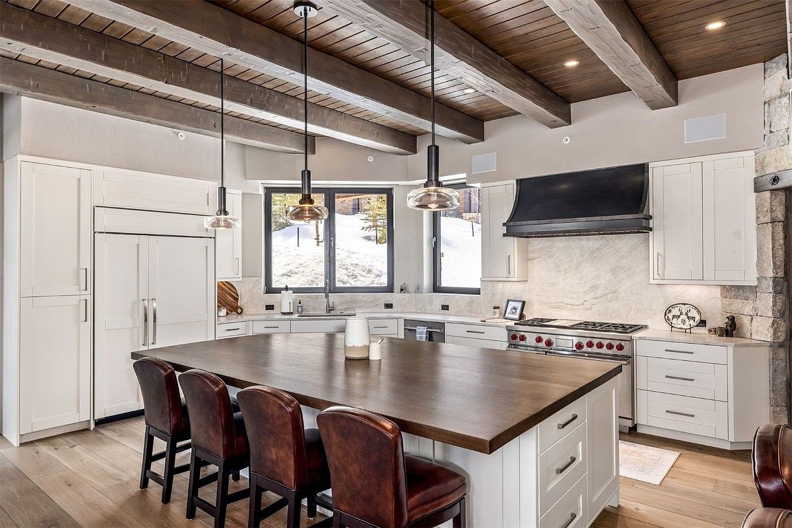 This is a well-lit kitchen featuring white cabinetry, a large dark wood island with seating, and stainless steel appliances. The kitchen has a rustic-modern design with exposed wooden beams on the ceiling and pendant lighting above the island. Large windows provide natural light and a view of the snowy landscape outside.