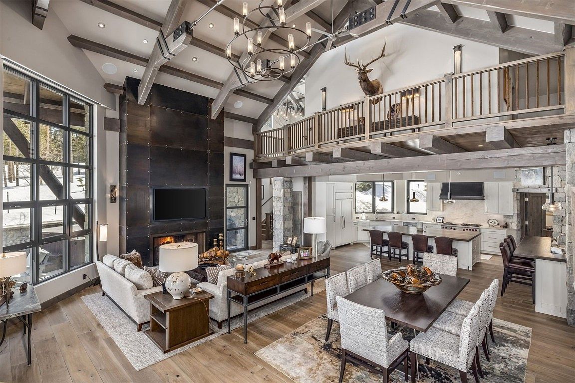 This is an interior shot of a luxurious living room that seamlessly blends into the kitchen and dining area. The living room features a large window, a modern fireplace with a dark metal surround, and comfortable seating. The open floor plan, exposed beams, and high ceilings create a spacious and inviting atmosphere, perfect for entertaining or relaxing.