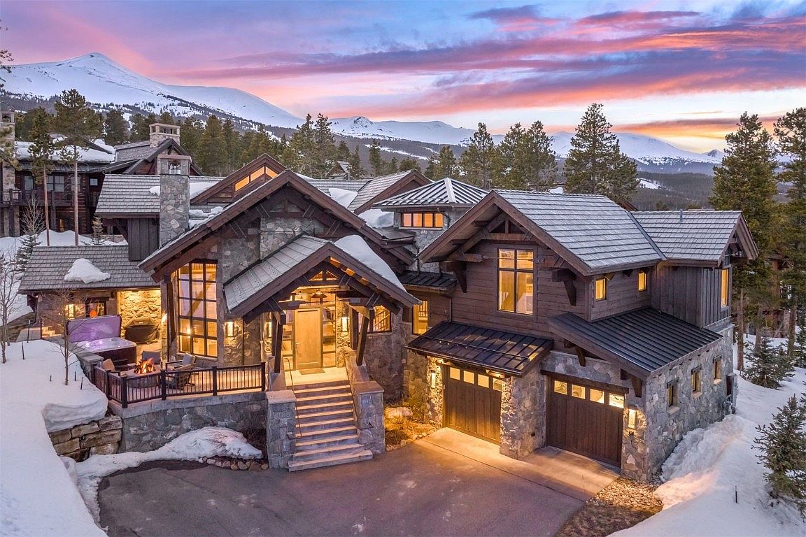 This is a stunning front view of a luxurious mountain home. The exterior features a combination of stone and wood, with a multi-level roofline and a grand entryway. A driveway leads up to a two-car garage, and snow-covered landscaping adds to the picturesque setting, with mountains visible in the background under a colorful sky.