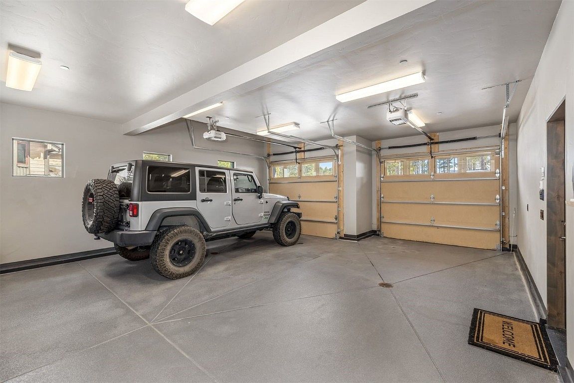 The image showcases a well-lit and spacious garage featuring two light-colored garage doors with windows, a gray epoxy floor with geometric patterns, and a gray Jeep Wrangler parked inside. The walls are painted in a neutral tone, and a small window provides natural light. A 'Welcome' mat is placed near the interior door, adding a touch of hospitality.