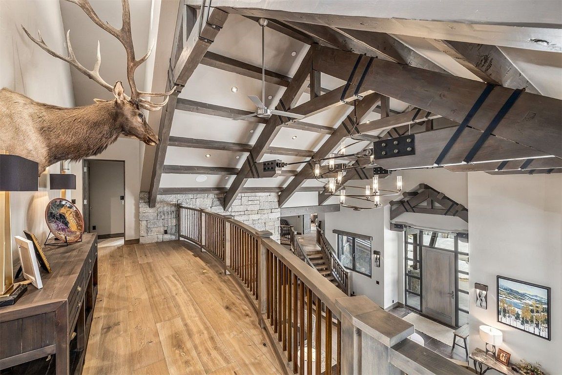 This interior shot showcases a luxurious hallway and staircase area. The space features hardwood flooring, a wooden railing, and exposed wooden beams on the ceiling, creating a rustic yet elegant atmosphere. A large mounted elk head adds a unique focal point, while the entryway below is visible, highlighting the home's grand scale and architectural details.