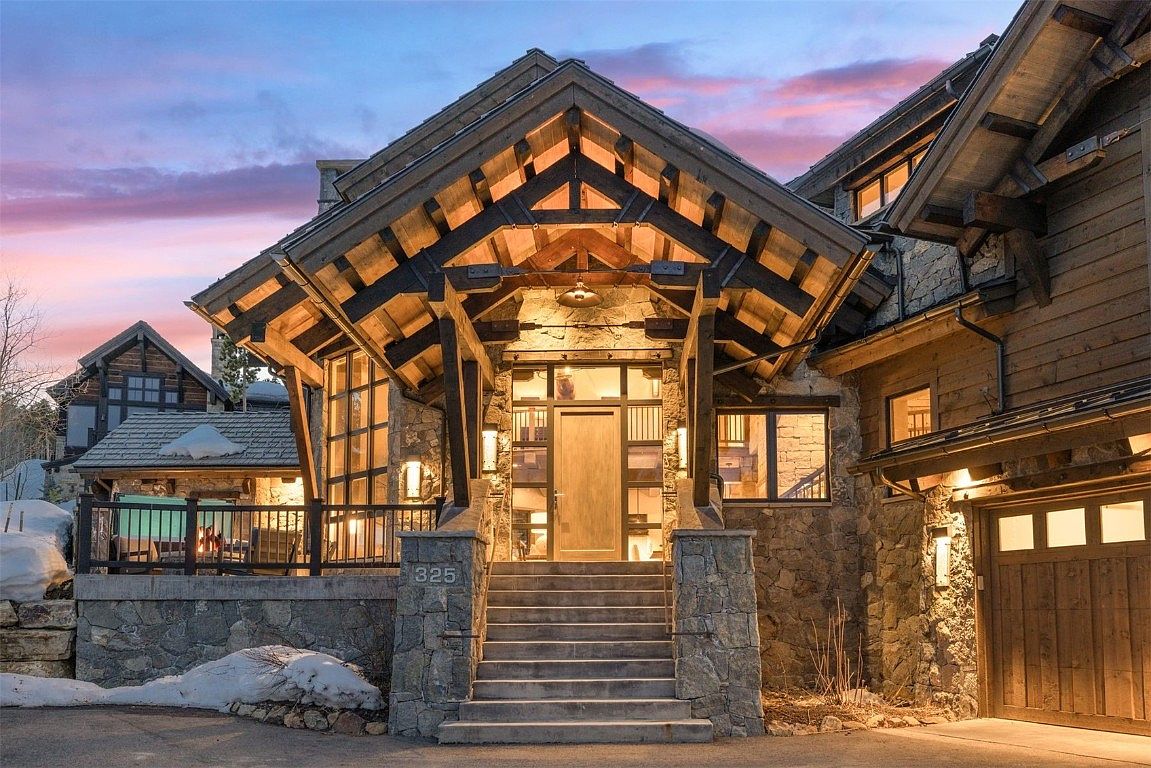 This is a captivating front entryway of a luxurious mountain home, showcasing a grand stone facade and a striking timber-framed porch. The entrance features a set of stairs leading to a wooden door framed by glass panels, with warm lighting accentuating the architectural details and creating an inviting ambiance. The property exudes a sense of rustic elegance and high-end craftsmanship.