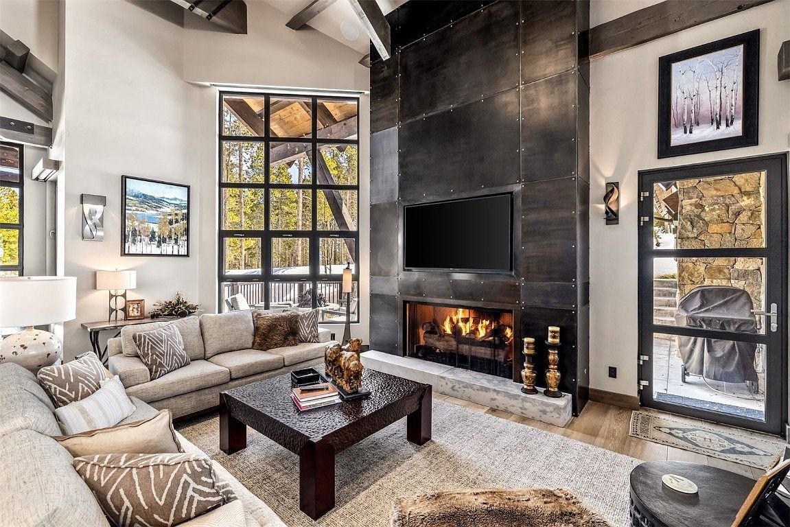 This is an interior shot of a luxurious living room featuring a stone fireplace with a mounted television. The room is furnished with comfortable sofas, decorative pillows, and a dark wood coffee table. Large windows offer natural light and views of the surrounding landscape, enhancing the room's cozy and inviting atmosphere.