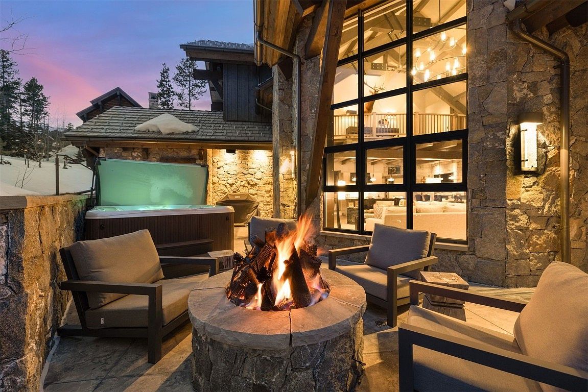 This image showcases a luxurious outdoor patio area, featuring a stone fire pit surrounded by comfortable seating. A hot tub is visible in the background, suggesting a relaxing and private space. The architecture incorporates natural stone elements, and large windows offer a glimpse into the interior, creating a seamless indoor-outdoor living experience.