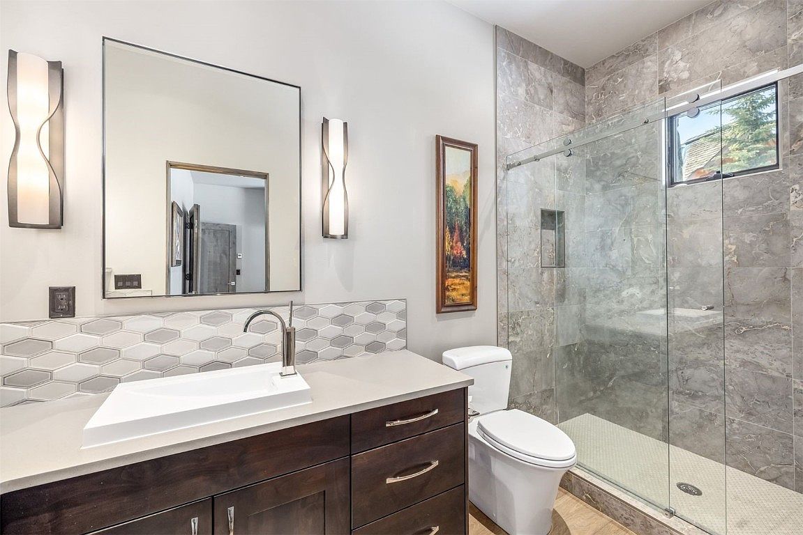 This is a well-lit bathroom featuring a modern design with a dark wood vanity, a white rectangular sink, and a sleek faucet. A large mirror hangs above the vanity, flanked by contemporary sconces. The shower area is enclosed with glass and features gray marble-like tiles, while the toilet sits adjacent to the vanity, creating a functional and stylish space.
