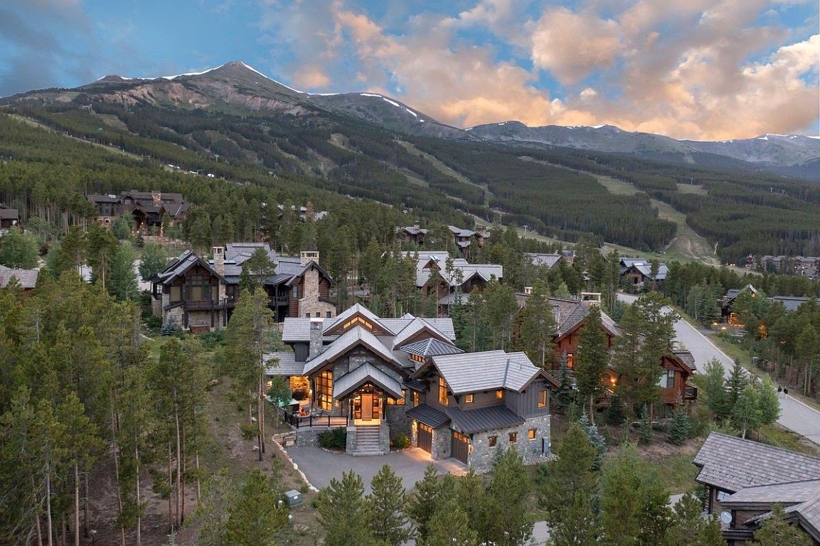 This aerial view showcases a luxurious mountain home nestled among tall pine trees, with a backdrop of majestic mountains partially covered in snow. The house features a stone and wood exterior, a multi-gabled roof, and a well-manicured driveway, exuding a sense of privacy and upscale living within a serene natural environment. The surrounding neighborhood consists of similar high-end homes, suggesting a prestigious community.