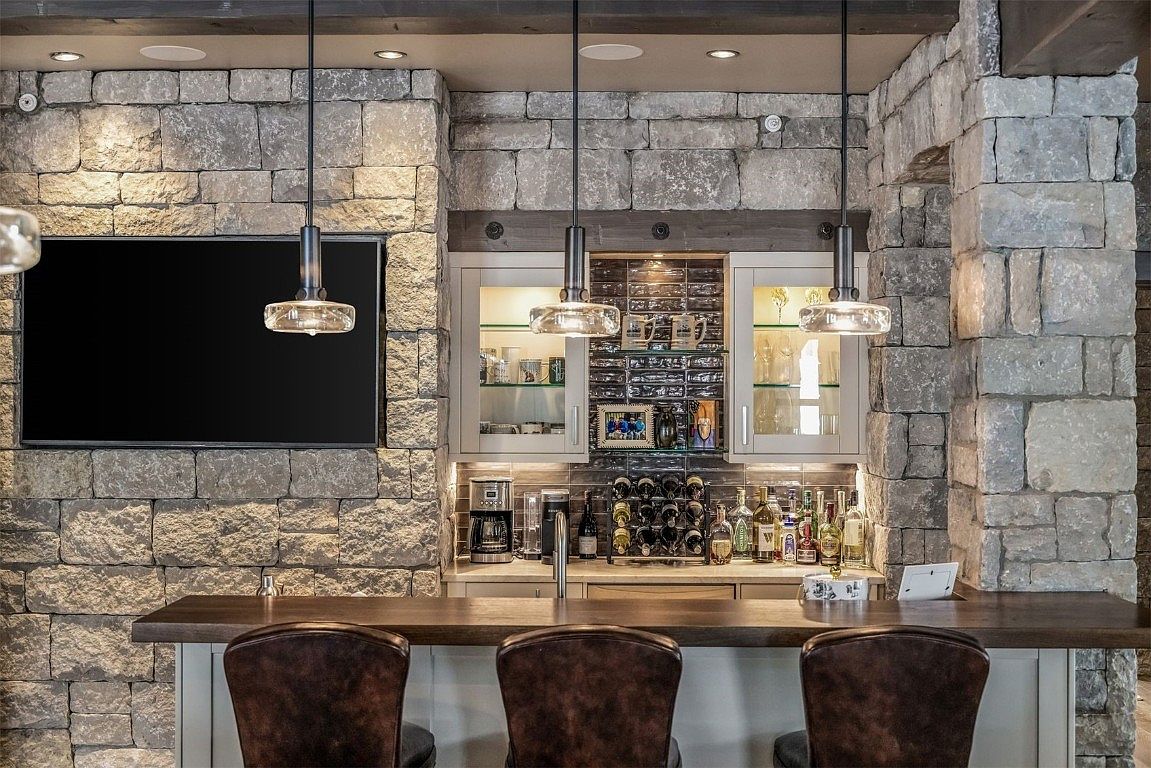 This interior shot showcases a sophisticated wine cellar or bar area, featuring a stone wall backdrop and a dark wood countertop. The space includes built-in cabinetry with glass doors, a wine rack, and various bottles and glassware, illuminated by pendant lighting. The overall impression is one of rustic elegance and refined taste.