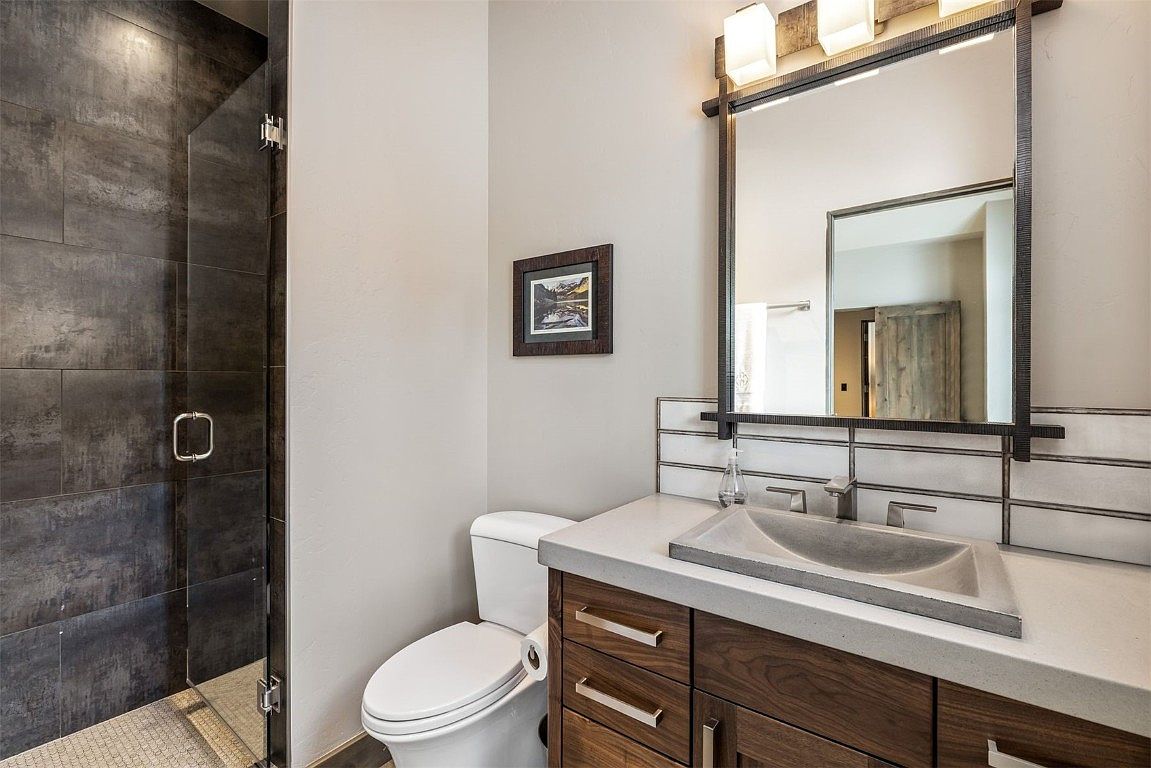 This is a well-designed guest bathroom featuring a modern aesthetic. The space includes a shower with dark, textured tiles and a glass door, a toilet, and a vanity with a concrete sink and wood cabinetry. A framed picture hangs on the wall, and a large mirror with integrated lighting adds to the contemporary feel.