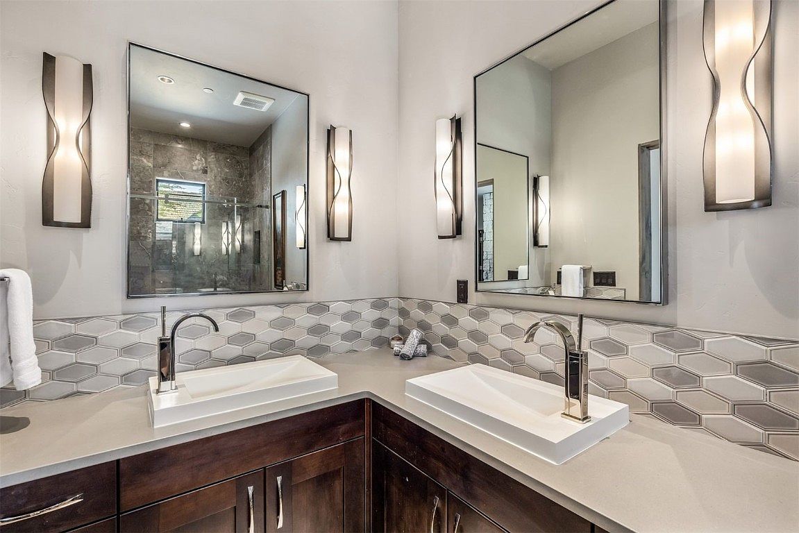 This is a well-lit primary bathroom featuring a double vanity with modern, rectangular sinks and sleek faucets. The backsplash is a stylish geometric tile pattern, and the mirrors are framed in black, complemented by unique sconce lighting. The dark wood cabinetry adds a touch of warmth to the contemporary design.