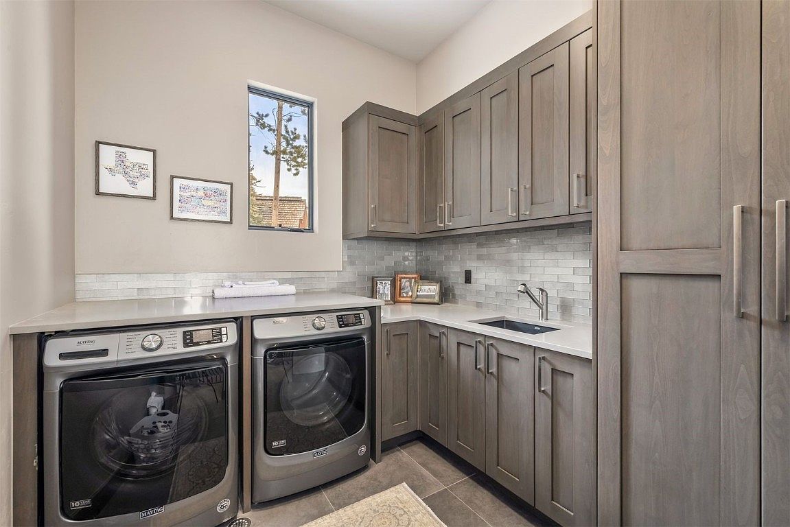 This is a well-organized laundry room featuring gray cabinetry with modern hardware, a white countertop, and a gray tile backsplash. A front-loading washer and dryer are neatly installed under the counter, and a stainless steel sink is integrated into the countertop. The room is well-lit and appears clean and functional, enhancing the home's appeal.