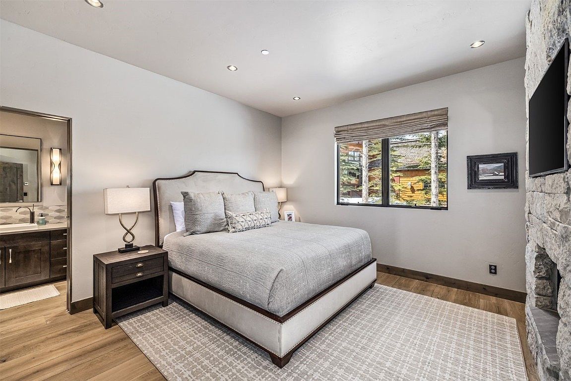This is a primary bedroom featuring a king-sized bed with a light-colored upholstered headboard and neutral bedding. A dark wood nightstand with a lamp sits beside the bed, and a window with a woven shade provides natural light. The room has a calm and inviting atmosphere, enhanced by the neutral color palette and soft textures.